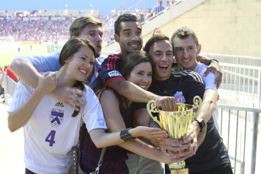 Sechs Menschen, zwei Frauen und vier Männer, die einen Pokal halten und lächeln, posieren für ein Foto vor einem Eisenzaun, mit einer Wand und einem Zaun oben rechts, Menschen und Lichter im Hintergrund, möglicherweise in einem Stadion.