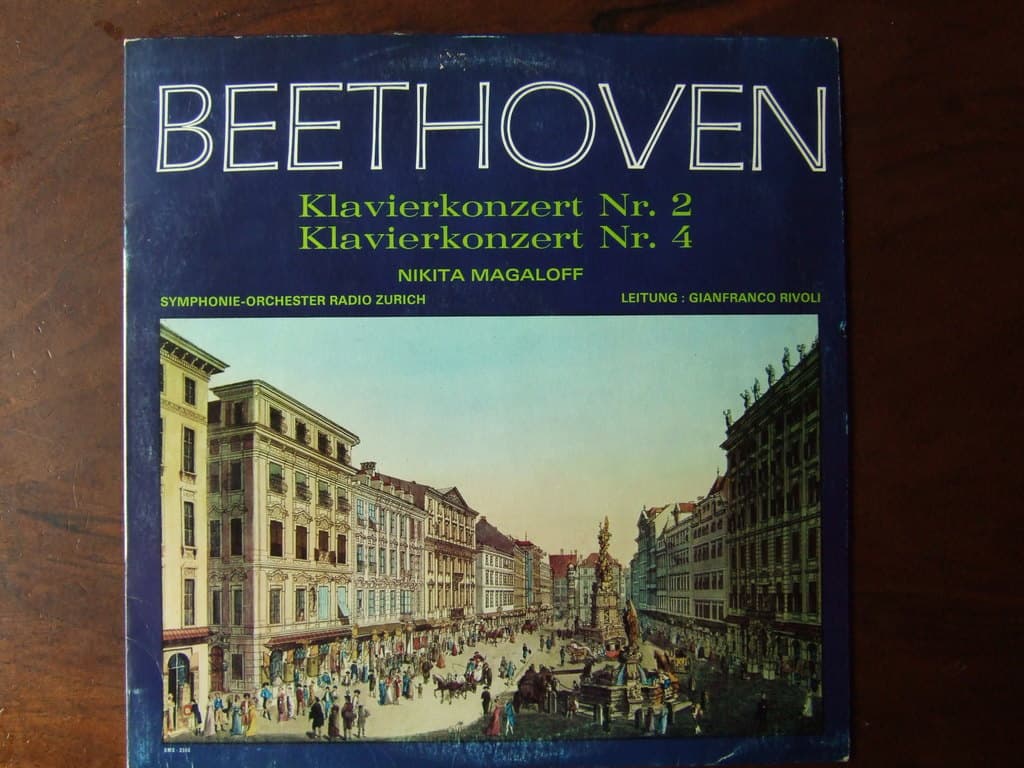 Ein Buch auf einer hölzernen Oberfläche zeigt ein Bild einer Straßen Szene mit vielen Menschen, Gebäuden auf beiden Seiten, einer Skulptur in der Mitte und dem Himmel darüber.