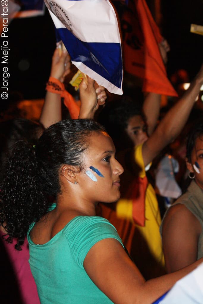 Eine Frau in einem gr√ľnen T-Shirt steht auf einer Demonstration und h√∂lt ein weißes Schild, hinter ihr stehen viele andere Menschen.