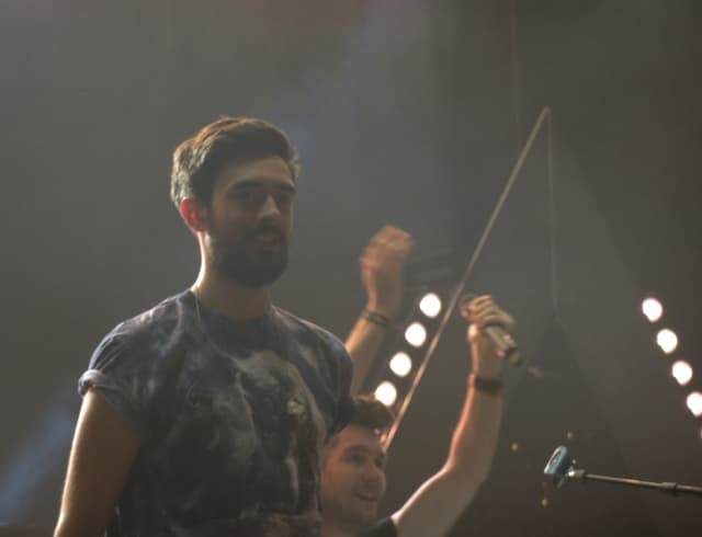 Ein Mann in einem T-Shirt steht und lacht, mit einem anderen lachenden Mann, Scheinwerfern, Rauch, einem Mikrofon und anderem Equipment im Hintergrund.