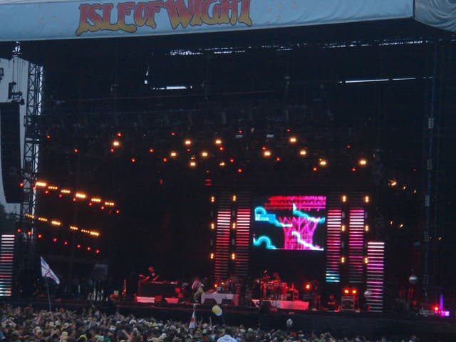 Eine Bühne mit Lichtern, einem Bildschirm und Musikern, die Instrumente spielen, mit einem Banner oben, einer Menge unten und Fahnen mit Stangen und Ballons drumherum.