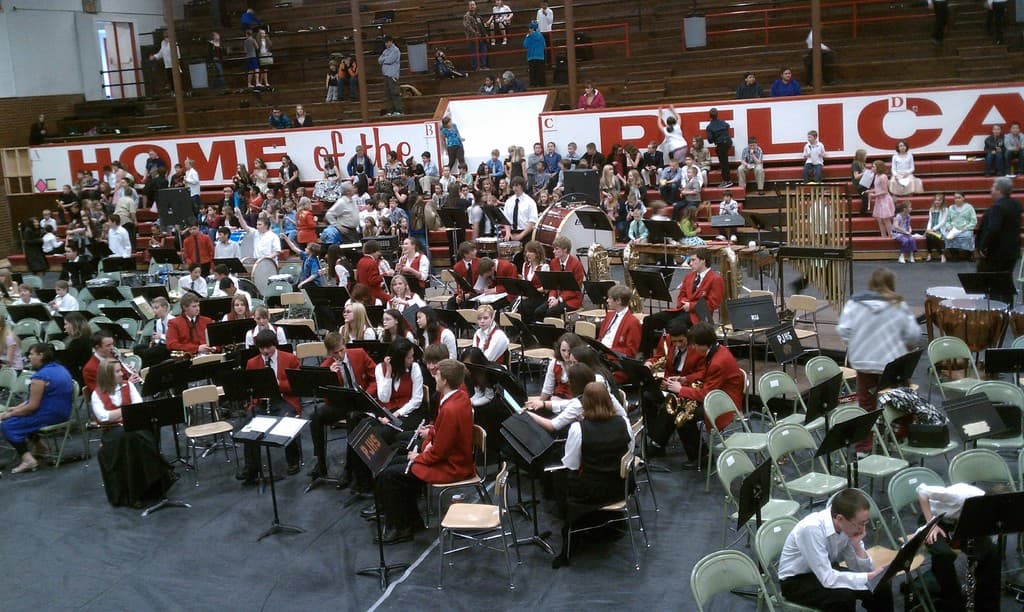 Eine Gruppe von Kindern sitzt auf Stühlen in einem Stadion während eines Musikunterrichts, mit Stadion-Sitzen im Hintergrund.