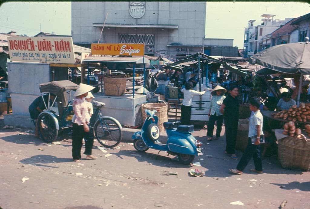 Eine belebte Stadtstraßenszene mit Geschäften, Zelten, Schirmen, einem Auto-Rikscha, einem Motorrad, Menschen und verstreuten Papiermüll in der Vordergrund, mit Gebäuden im Hintergrund.