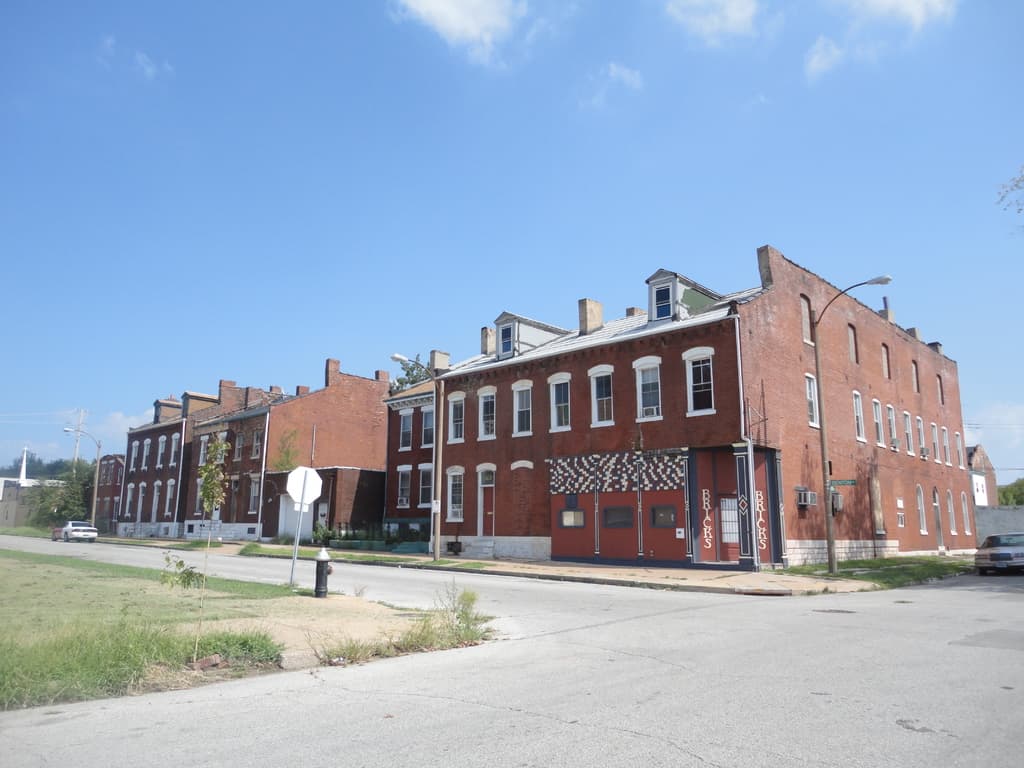 Ein Gebäude mit Masten, Straßenlaternen und Bäumen, Fahrzeuge auf beiden Seiten, ein Gehweg und Gras unten, ein Feuerlöscher und der Himmel oben.