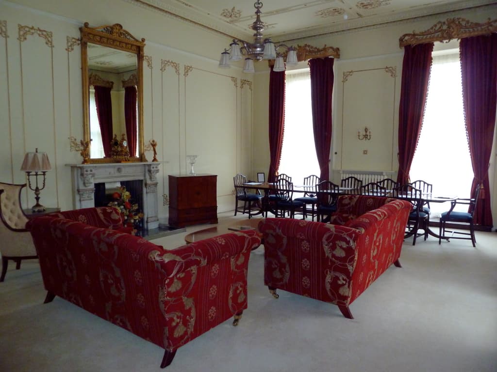 Zwei rote Sofas in einem Wohnzimmer, ein kleiner Teetisch mit einem Buch darauf, ein wandmontierter Spiegel, eine Lampe auf einem Tisch, ein Blumenbouquet vor einem Kamin, ein Esstisch mit Stühlen, von der Decke hängende Deckenleuchten, Vorhänge an einer Stange und ein leerer Stuhl.