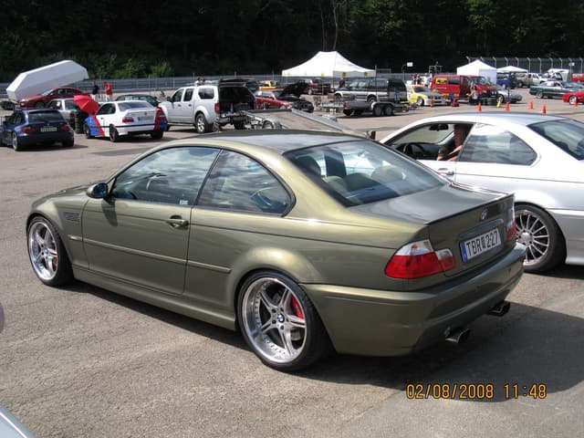 Eine Au├čenaufnahme mit mehreren Fahrzeugen auf einem Parkplatz, einem weißen Zelt in der Ferne, zahlreichen B├Ąumen, einigen Menschen, die stehen, einem roten Schirm und zwei Autos im Vordergrund.