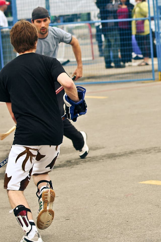 Zwei Menschen spielen Hockey auf einer Straße mit einem Zaun dahinter und einer Menge Menschen im Hintergrund.