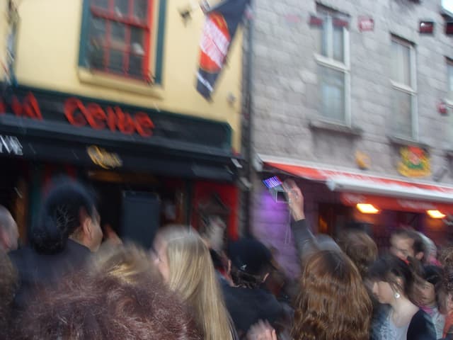 Ein unscharfer Bildausschnitt, der unten Menschen auf der Straße und oben Gebäude mit Lichtern, Fenstern und einer Fahne zeigt.