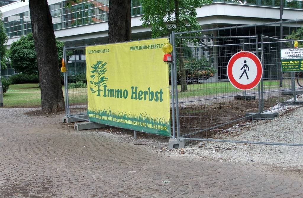 Ein Zaun mit einer Fahne, Bäumen und Pflanzen links, ein Gebäude im Hintergrund und ein Kopfsteinpflasterweg unten.