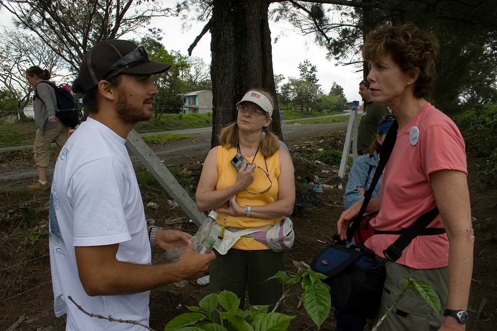 Eine Frau in der Mitte hält eine Kamera, ein Mann links hält eine Flasche, mit einem Himmel und Bäumen im Hintergrund.