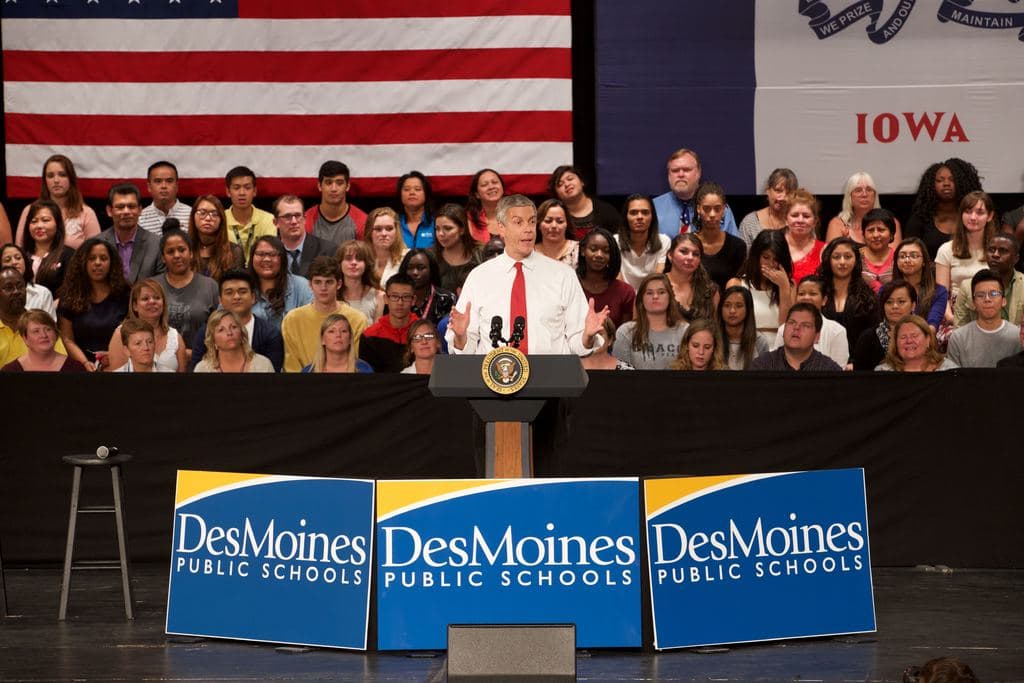 Ein Mann in weißem Hemd und roter Krawatte steht an einem Rednerpult mit drei Tafeln, einem Hocker mit Mikrofon und einer sitzenden Menge im Hintergrund, mit einer Flagge und zusätzlichem Text.