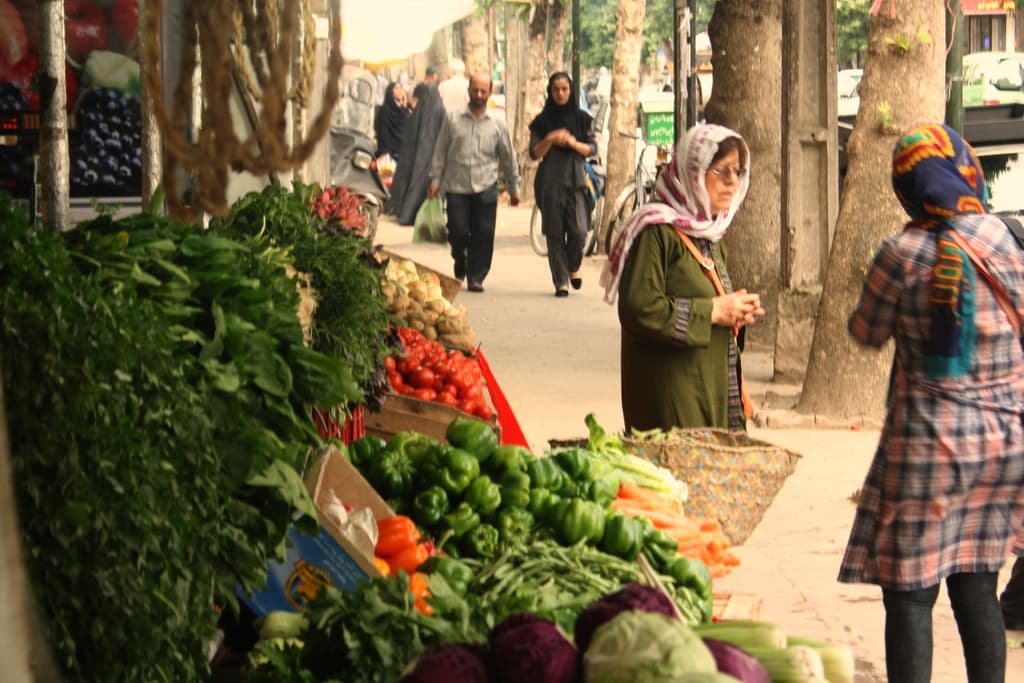 Ein belebter Gemüsemarkt mit verschiedenen Produkten wie Paprika, Tomaten und Kartoffeln auf der linken Seite, Menschen, die stehen und gehen, auf der rechten Seite und Bäume und Gebäude im Hintergrund.