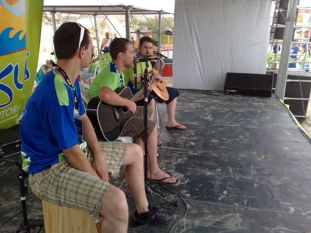 Drei Männer sitzen auf Stühlen auf einer Bühne, zwei spielen Gitarren, mit einem Mikrofonständer davor, einem Banner dahinter und einem schwarzen Kasten auf der rechten Seite der Bühne, mit einer Hütte, Menschen, Bäumen und Gebäuden im Hintergrund.