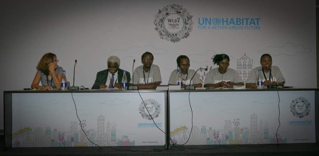 Sechs Personen in einem Konferenzsaal, eine Frau in einem grauen Kleid auf der linken Seite, eine Flasche auf dem rechten Tisch, ein weißes Banner im Hintergrund und zwei Tische mit Büchern, Mikrofonen und Flaschen im Vordergrund.