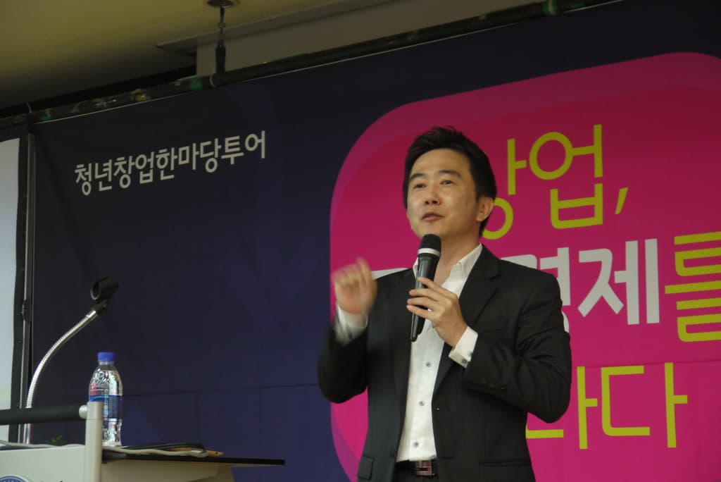 Eine Person in einem schwarzen Anzug und weißem Hemd hält ein Mikrofon und steht neben einem Tisch mit einer Flasche darauf, mit einem Plakat mit Text dahinter.