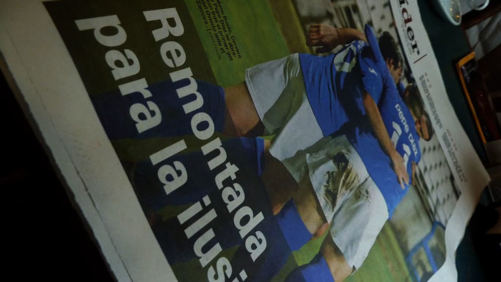 Zeitung auf einem Tisch zeigt drei Männer in blauen T-Shirts, möglicherweise beim Fussballspielen, mit Text unten im Bild.