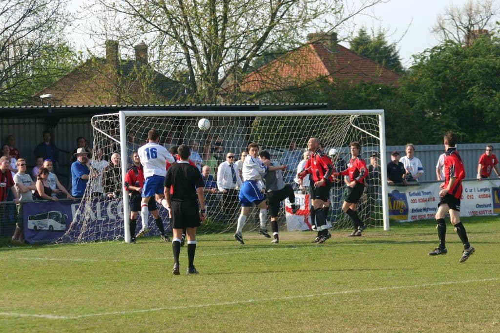 Players are engaged in a football game on a field with a goal net, while spectators stand behind it, with trees and houses visible in the background.