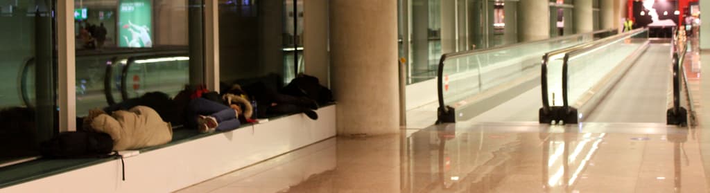 Menschen auf Bänken in einem Flughafen mit zwei geraden Rolltreppen im Hintergrund.