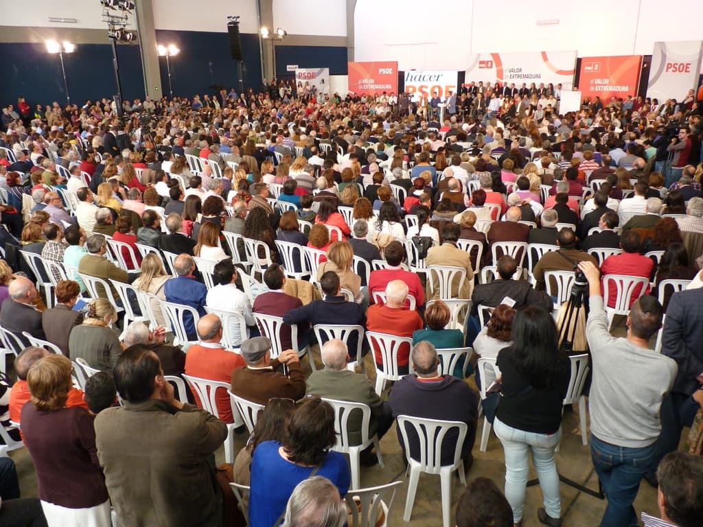 Ein volles Stadion mit vielen sitzenden und stehenden Menschen, eine Bühne mit Personen in der Nähe eines Podiums, zwei sprechende Personen und verschiedene Banner und Lichter.