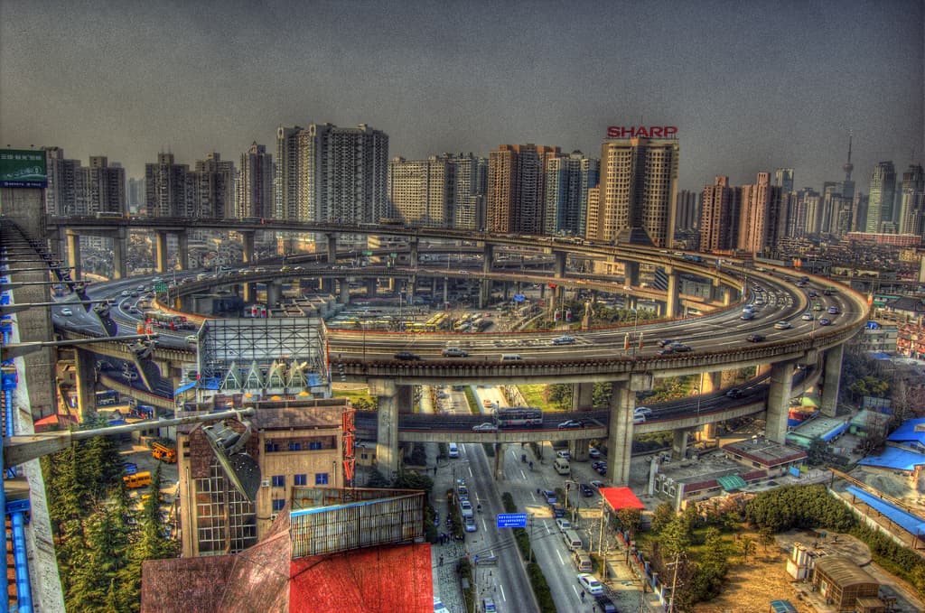 Eine Stadtansicht mit einer zentralen Ringstraße mit Fahrzeugen, einer unteren Straße mit Autos und Fahrrädern, zahlreichen Hochhäusern im Hintergrund, einem klaren Himmel darüber, Bäumen auf der linken unteren Seite und kleinen Häusern, die hier und da in der Stadt verteilt sind.