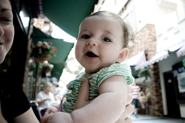 Eine Frau mit einem Kind auf dem Arm vor einem unscharfen Hintergrund mit einer sitzenden Frau, einem gehenden Mann, Hauspflanzen, Gebäuden und verschiedenen Gegenständen.