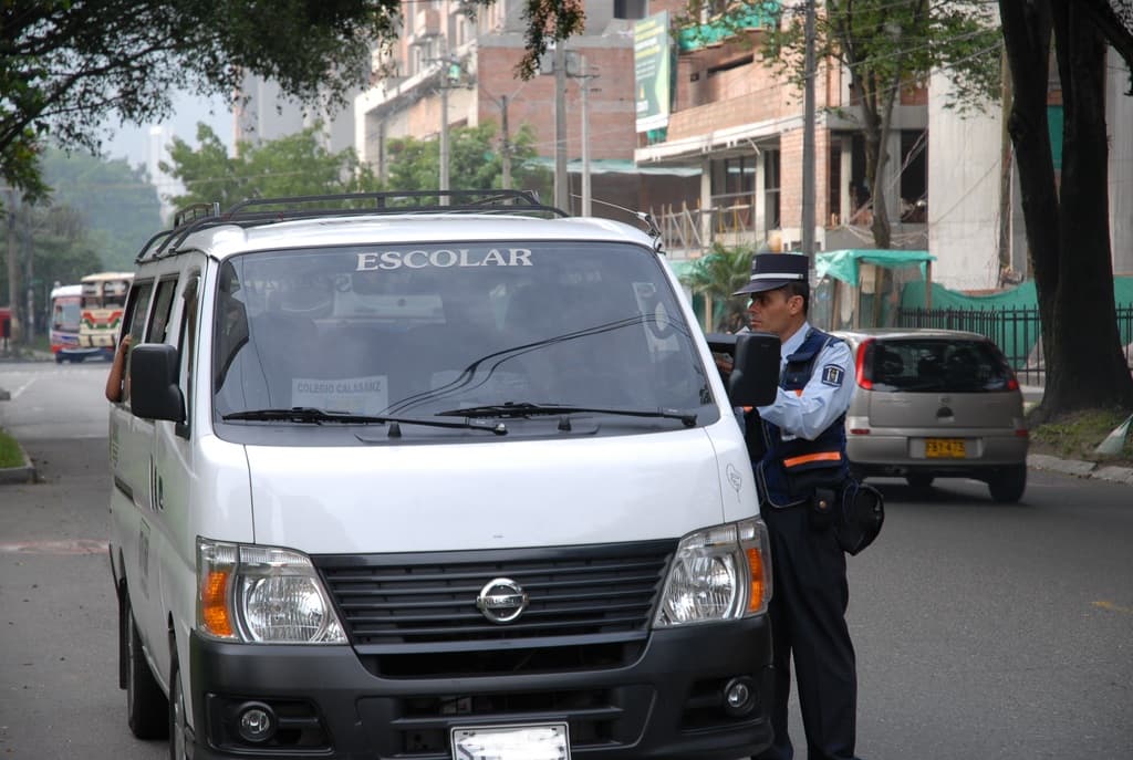 Ein Polizist spricht mit einer Person in einem Fahrzeug auf einer belebten Straße, die von Bäumen, hellbraunen Gebäuden und Strommasten gesäumt ist.