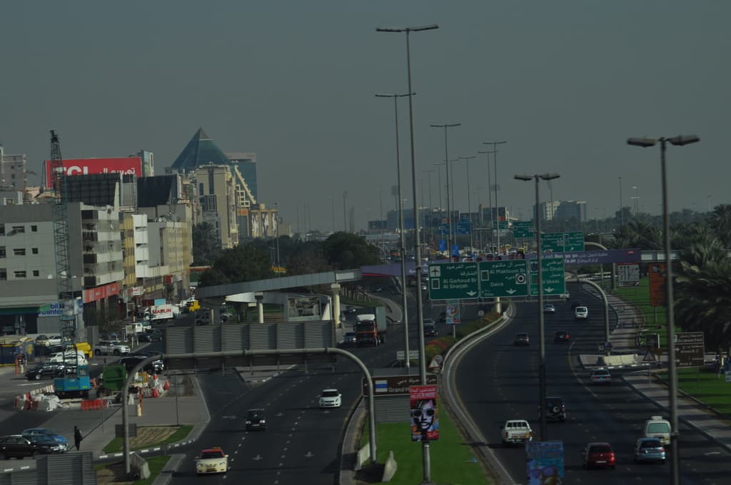 Eine Luftaufnahme einer Stadt mit Gebäuden, Türmen, Straßen voller Fahrzeuge, zahlreichen Werbetafeln und Schildern, Straßenlaternen, Bäumen, Menschen und Verkehrskegeln.
