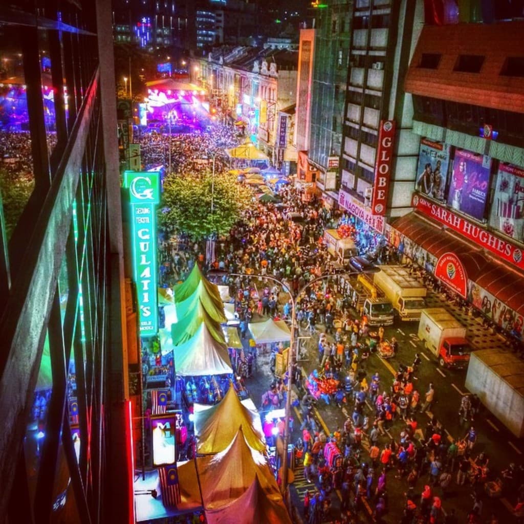 Ein volles Karnevalszelt mit Zelten, Ständen, Gebäuden und Bannern, mit beleuchteter Fläche im Hintergrund.