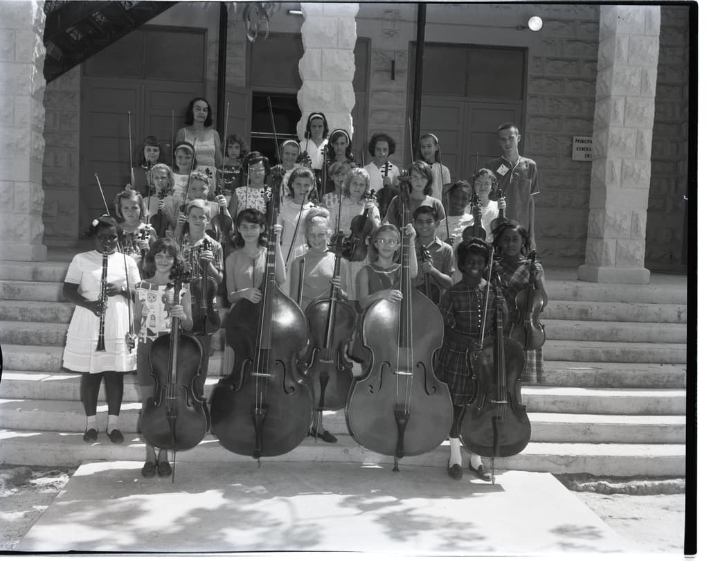 Kinder auf einer Treppe mit Musikinstrumenten wie Celli, Geigen und Trompeten, im Hintergrund Säulen und eine Wand mit Türen.