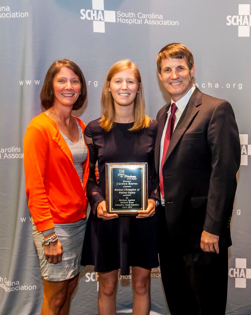 Drei Personen, zwei Frauen und ein Mann, stehen zusammen. Der Mann trägt einen schwarzen Anzug, eine Frau trägt ein orangefarbenes und schwarzes Kleid und die andere Frau trägt ein schwarzes Kleid und hält einen Preis in den Händen. Sie stehen vor einem grauen Hintergrund.