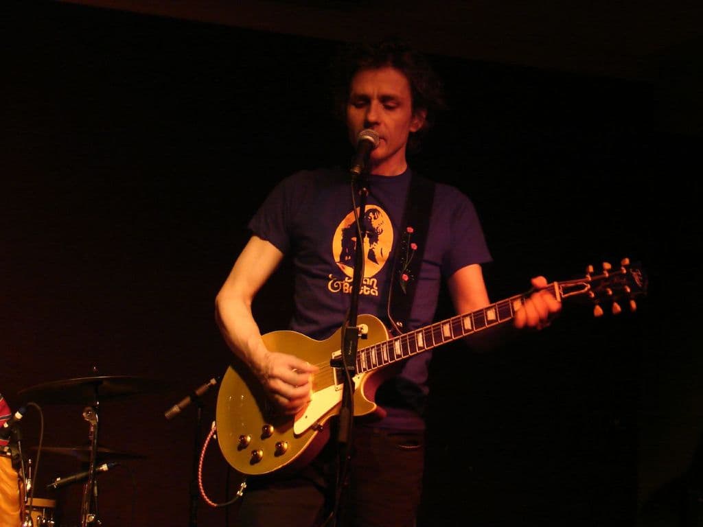 Eine Person steht mit einer Gitarre und singt in ein Mikrofon, während mehrere Musikinstrumente links zu sehen sind.