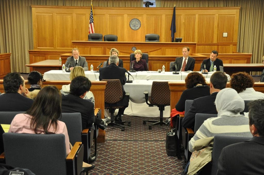 Ein Versammlungsraum mit einer Gruppe von Menschen, die auf Stühlen sitzen, zwei ausländische Flaggen, ein oranger Schreibtisch mit Wasserflaschen und einem Mikrofon.