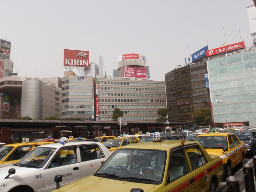 Eine Stadtansicht mit Fahrzeugen auf der Straße, Gebäuden, Bäumen, Pfählen mit Schildern und Text auf den Schildern, einem Zug im Hintergrund und einem Geländer rechts unten.