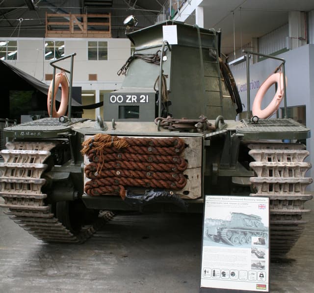 Ein Panzer-Panzer ist auf dem Boden positioniert mit einer Tafel und mehreren Bildern in der Nähe, gegen einen Hintergrund mit einer Wand, Säulen, Lampen, einem Schuppen und Stangen.