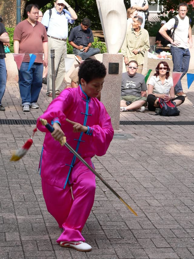 Eine Person in einem rosafarbenen Kleid hält ein Schwert, während viele Menschen zusehen.