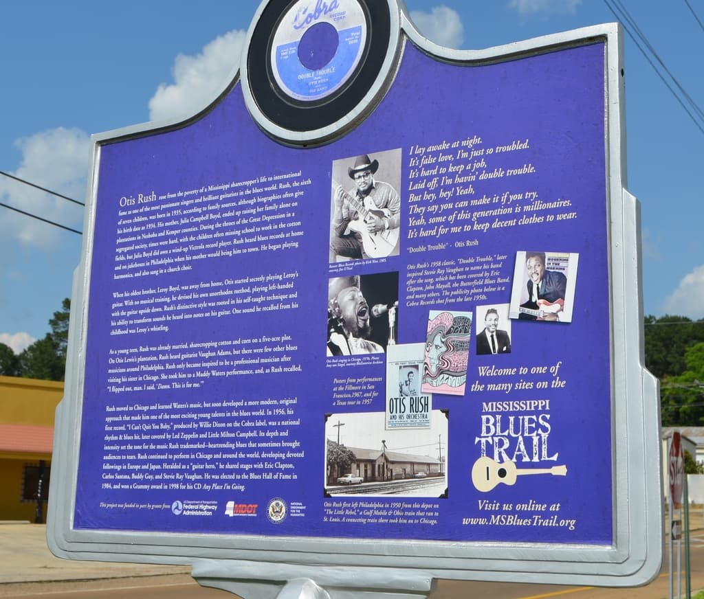 Ein Informationsschild, Gebäude, ein Schuppen, Bäume und Stromkabel unter einem bewölkten Himmel.