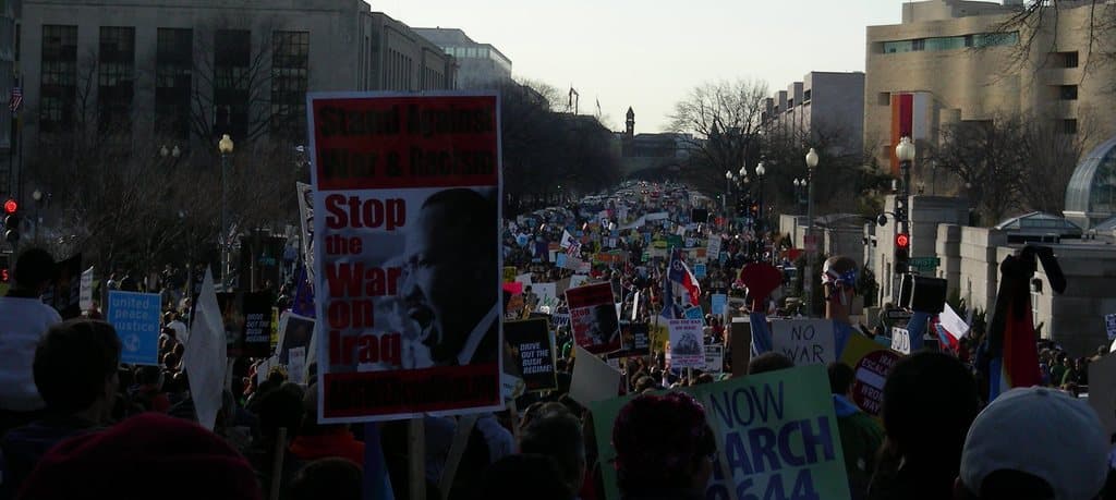 Menschen protestieren auf einer Straße und halten Plakate in Hönden, mit Gebäuden, Bäumen, Laternenmästen und dem Himmel im Hintergrund.