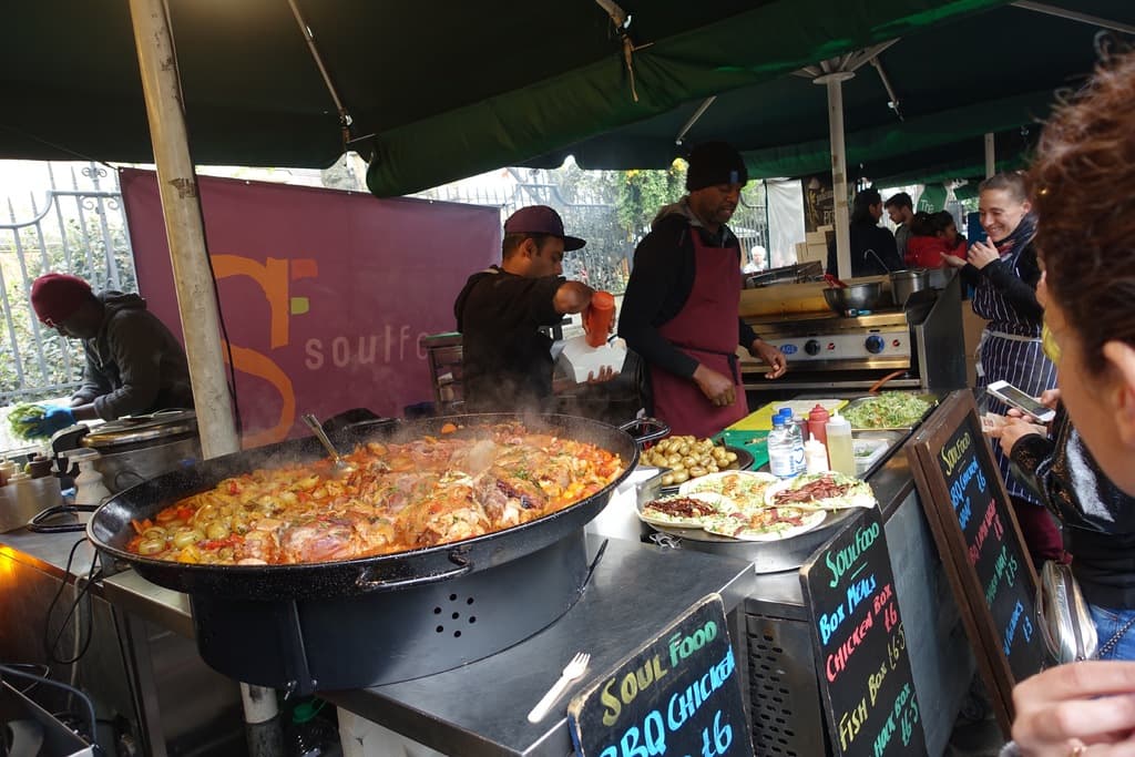 Ein Imbissstand mit einer Gruppe von Menschen, die stehen, eine Person in der Mitte hält eine Flasche auf einer Kiste, ein Herd rechts, eine Tafel unten rechts und Essen unten links.