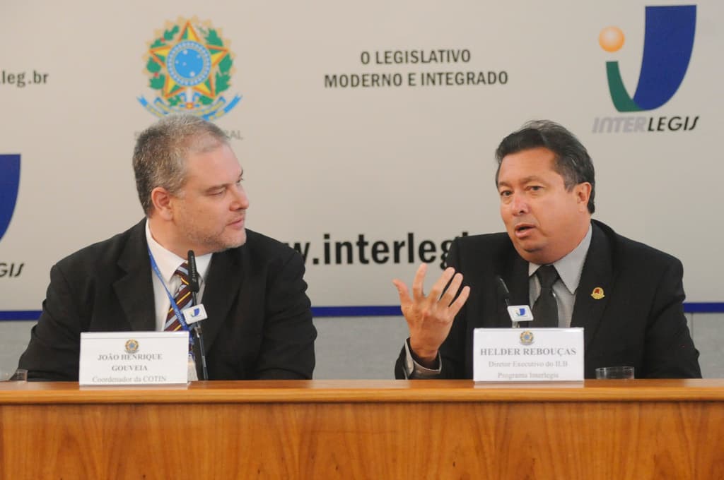 Zwei Männer in Anzügen sitzen an einem Tisch mit zwei Namensschildern und zwei Mikrofonen, mit Logos und Text im Hintergrund.