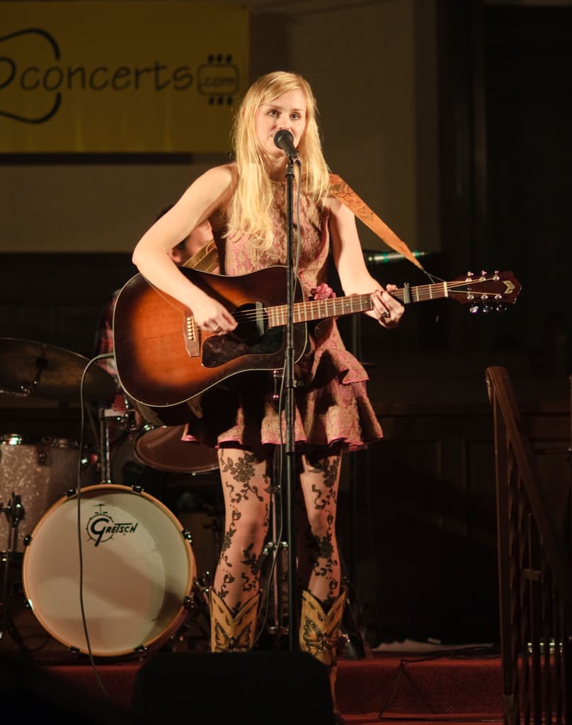 Eine Frau, die auf der Bühne eine Gitarre spielt und singt, mit einer Band im Hintergrund und einem Bildschirm an der Wand dahinter.