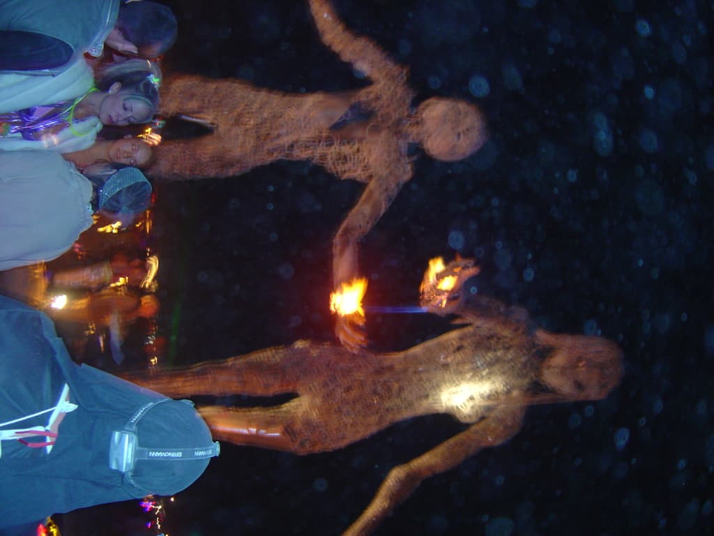 Eine Gruppe von Menschen, die vor brennenden Statuen mit Lichtern im Hintergrund stehen.