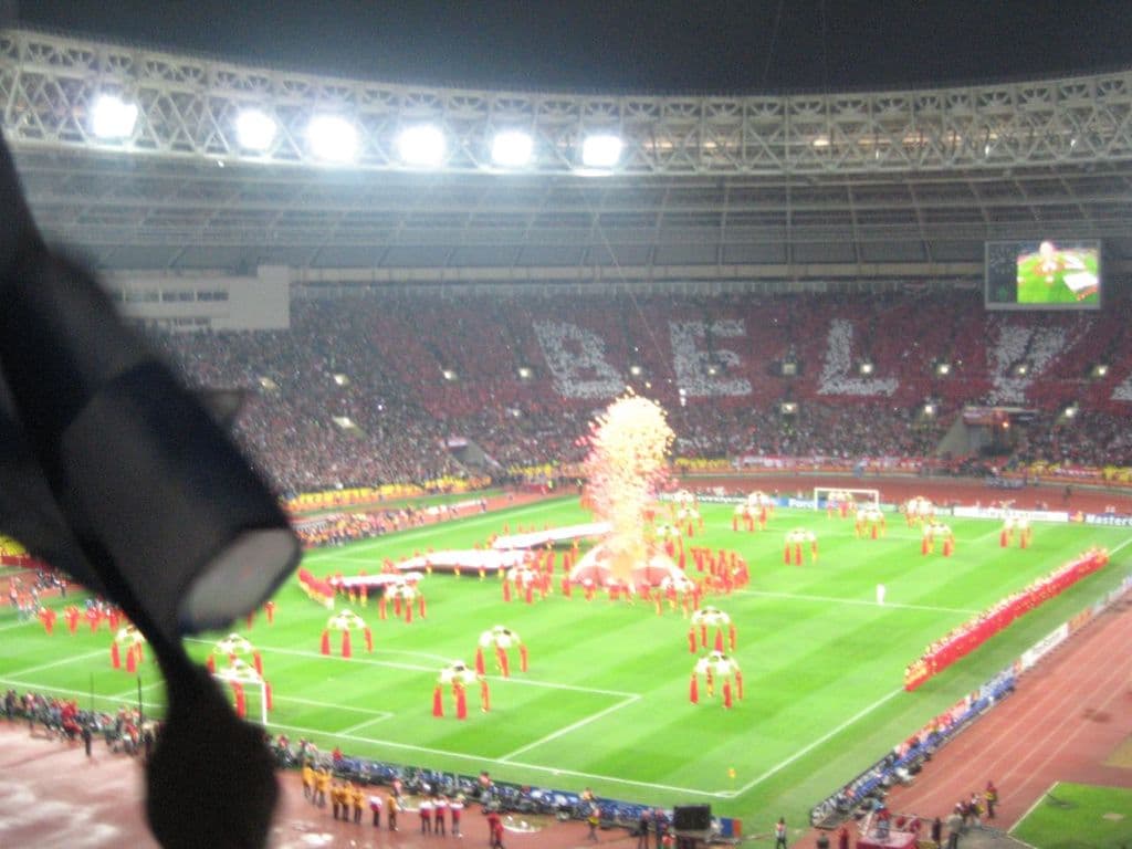 Ein Stadion mit Flutlicht, Eisengittern, Zuschauern, Menschen auf dem Boden, Werbetafeln, Anzeigetafeln, Überwachungskamera und Fahnen und Dekorationen.