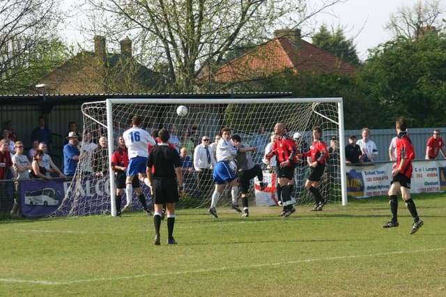 Spieler spielen Fußball auf einem Feld mit einem Tor, während Zuschauer dahinter stehen, mit Bäumen und Häusern im Hintergrund.