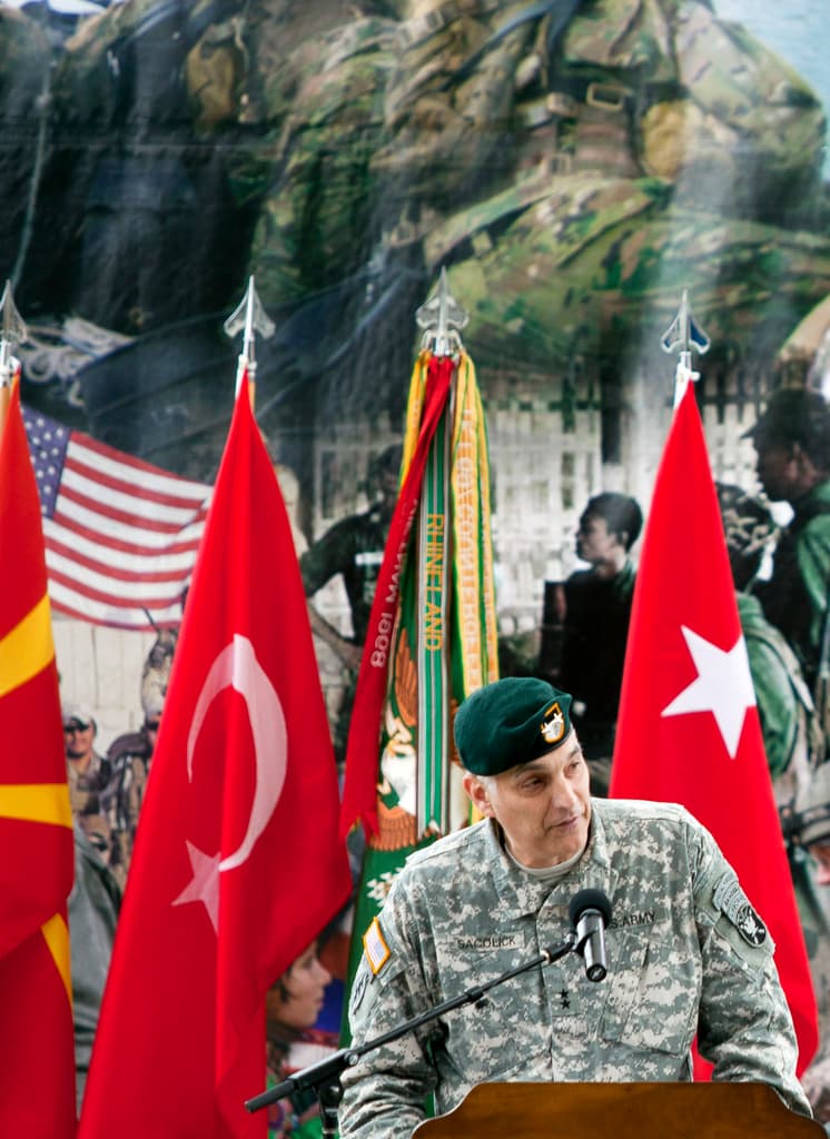 Ein Soldat steht auf einem Podium mit einem Mikrofon, mit verschiedenen Flaggen und einem Plakat mit Militärpersonal im Hintergrund.