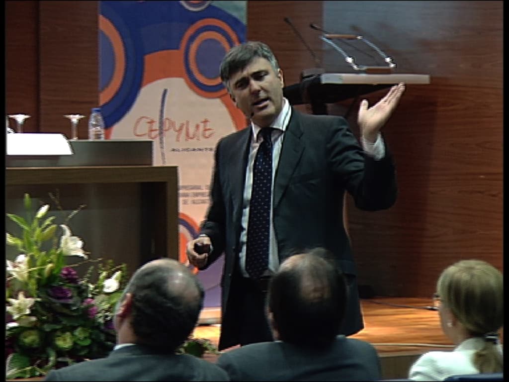 Ein Mann in einem schwarzen Blazer steht an einem Podium mit Mikrofonen und spricht in eines hinein, mit einem Blumentopf oder einem Bouquet in der Nähe und einem Tisch mit einem Buch, einer Wasserflasche und anderen Gegenständen daneben. Im Hintergrund ist eine braune Wand und eine farbenfrohe Tafel mit Text zu sehen, was auf eine Konferenzhalle hindeutet.