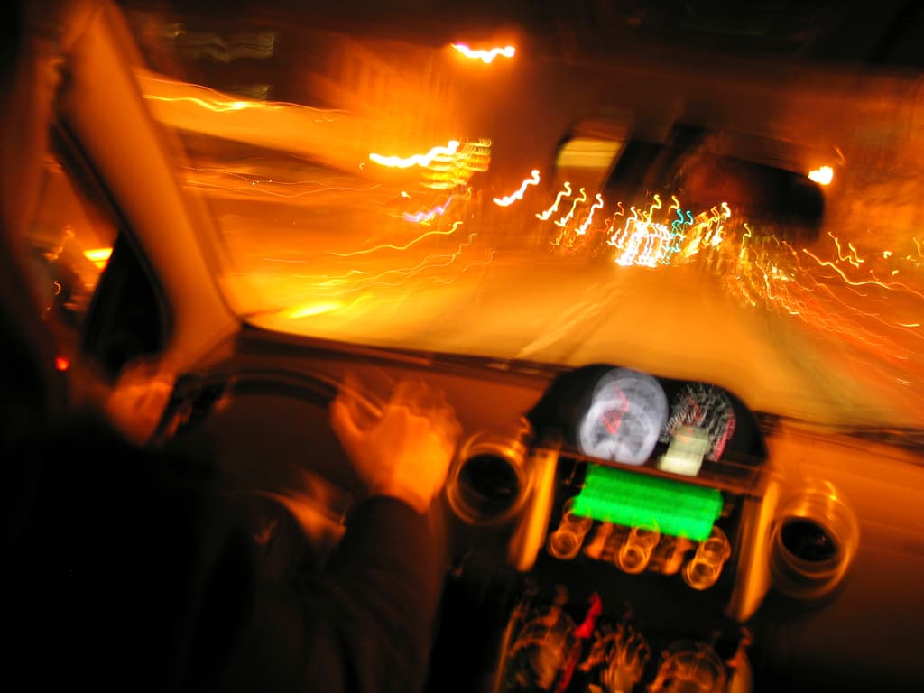 Eine Person sitzt in einem Auto und schaut durch eine Scheibe auf Lichter, eine Straße und Gebäude.