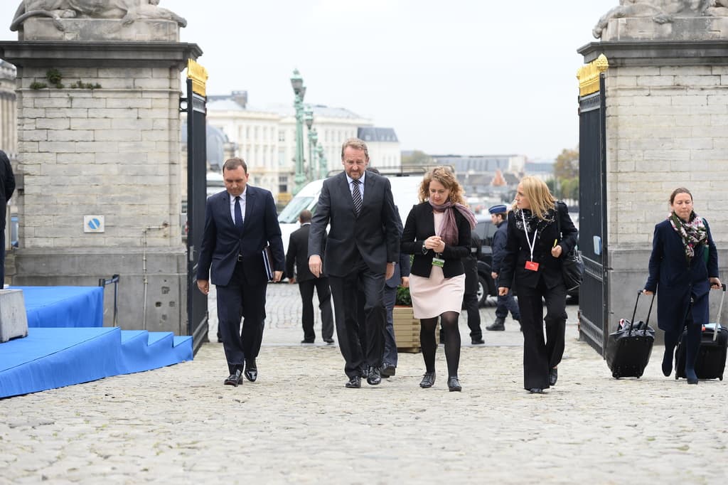 Eine Gruppe von Mitarbeitern geht in einen Bereich mit einem Tor, hinter dem sich viele Fahrzeuge und Gebäude befinden, während eine Frau auf der rechten Seite zwei Koffer trägt.
