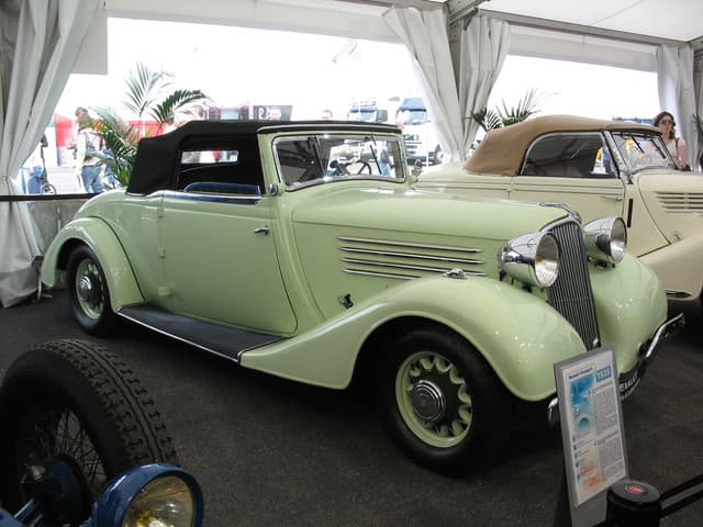 Mehrere Oldtimer stehen auf einem Boden mit Vorhängen und Pflanzen im Hintergrund, während weiter hinten auf der Straße einige Menschen und Fahrzeuge zu sehen sind.