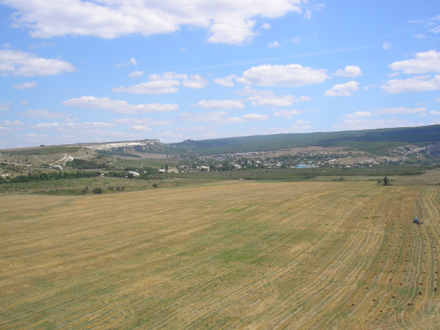 Ein Traktor, der auf einem kultivierten Feld mit Bäumen, Gebäuden, Hügeln und einem bewölkten Himmel im Hintergrund arbeitet.