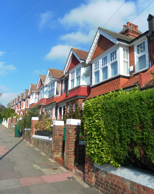 Eine Reihe von Reihenhäusern mit Glasfenstern auf einer Londoner Straße, umgeben von Bäumen und Pflanzen unter einem Himmel mit weißen Wolken.